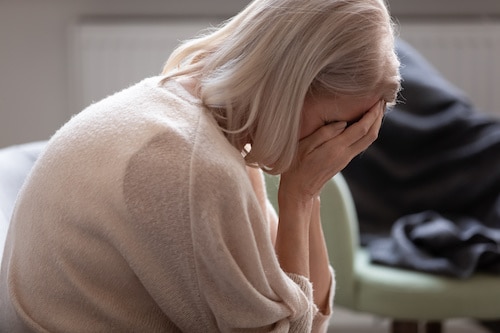 Senior woman hides face with hands crying seated on couch